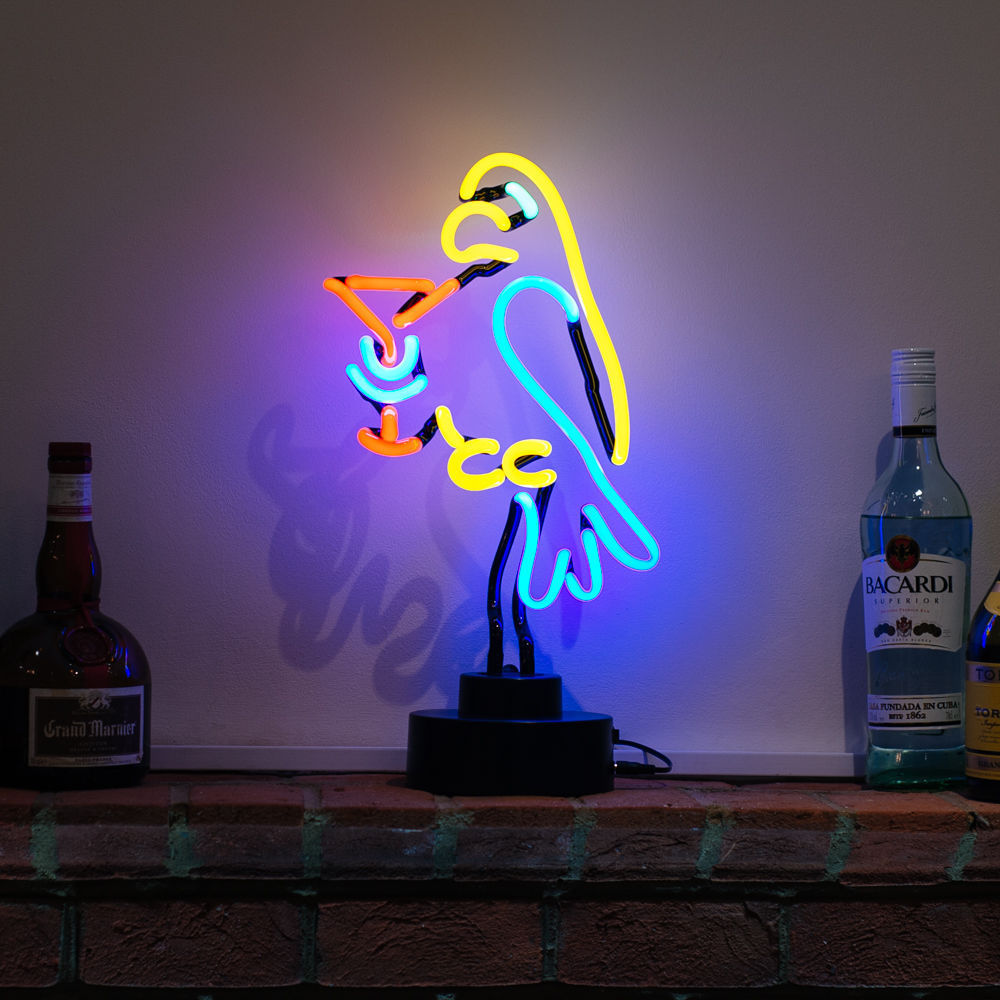 A table-top neon sign showing a colorful neon parrot and cocktail drink glowing red, blue, and yellow on a table against a blurred brick background.