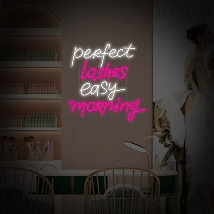 Close-up photograph of the "Perfect Lashes Easy Morning" neon sign, highlighting its elegant script and soft pink illumination against a textured wall.