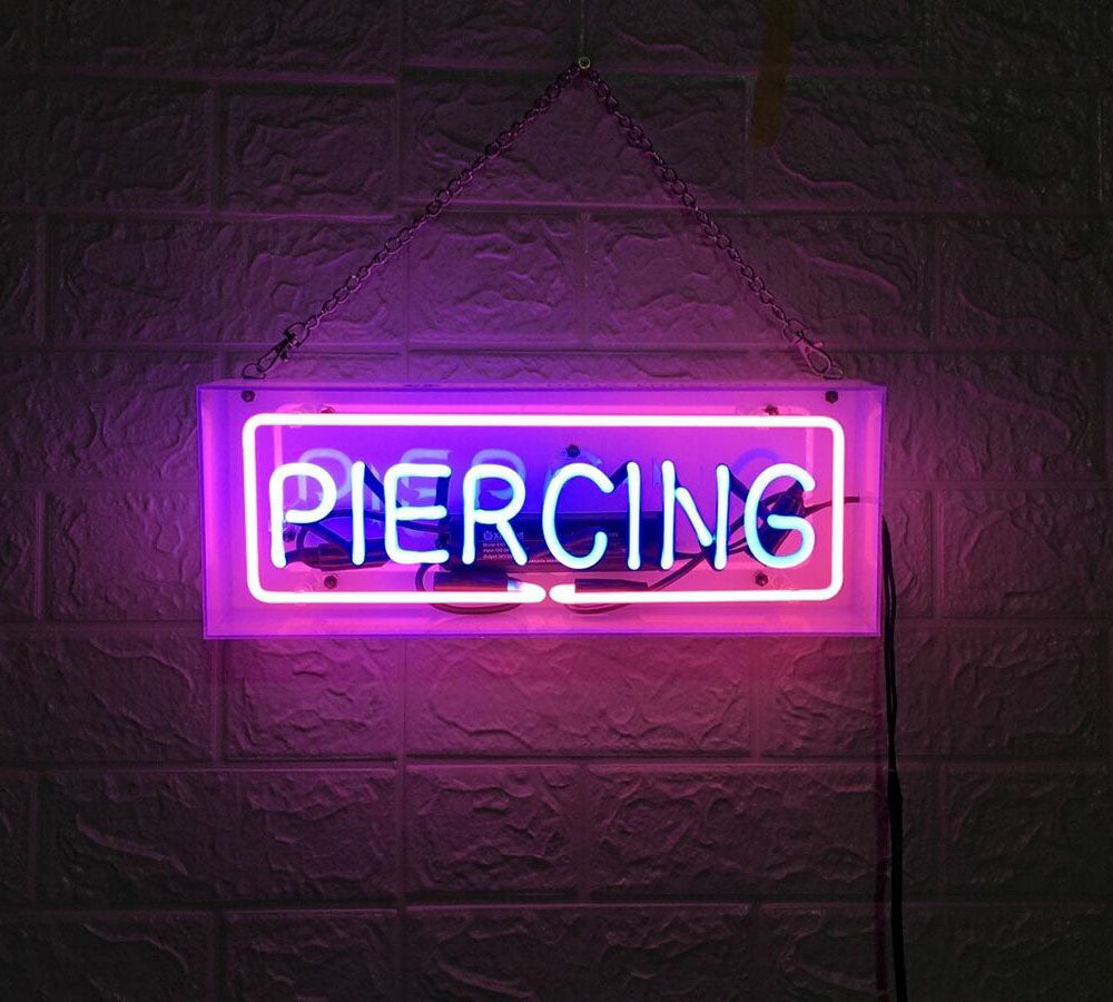 A close-up photograph of a blue and pink 'Piercing' neon sign encased in a clear acrylic box, showing realistic glass tubes and internal wiring against a textured wall.