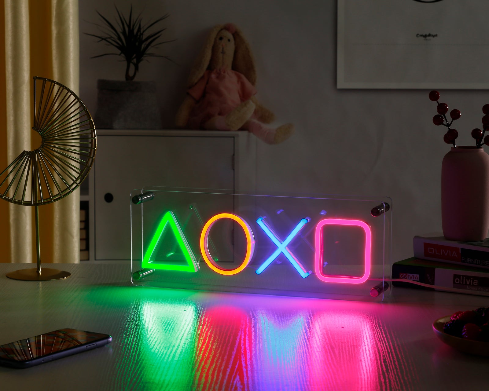 A close-up photograph of the illuminated Play Gaming Station Geometry desk neon sign with exposed hardware and wiring, glowing intensely in a dark, prop-filled environment.