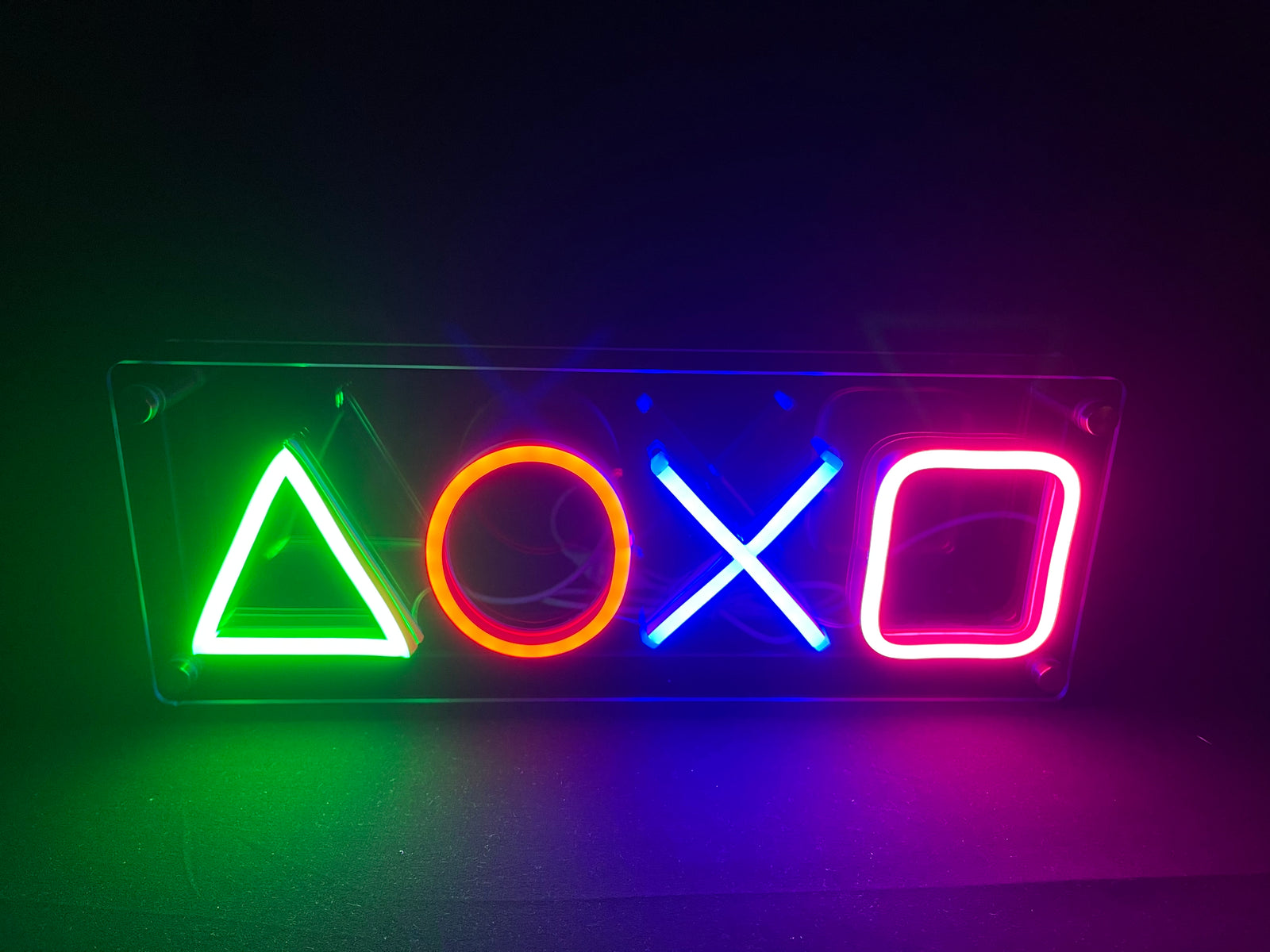 The front view of the glowing blue, red, green, and pink geometric symbol neon sign mounted on a clear acrylic backboard against a dark, slightly reflective surface.