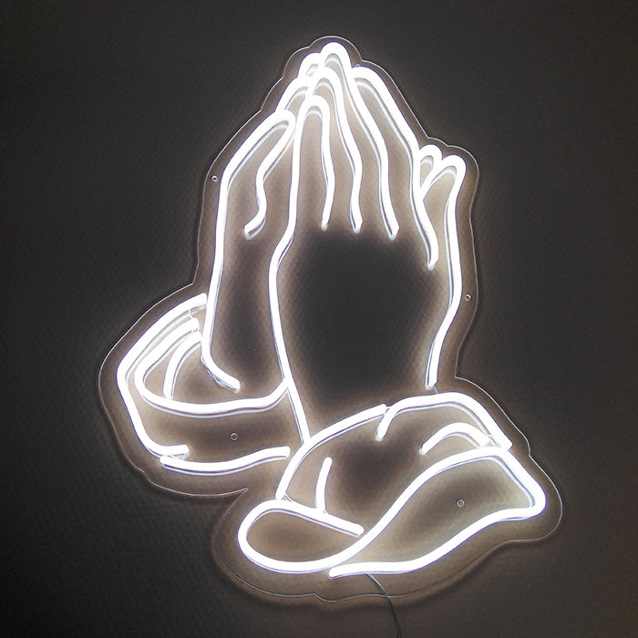 A glowing yellow neon sign depicting clasped praying hands mounted on a clear backboard against a neutral wall.