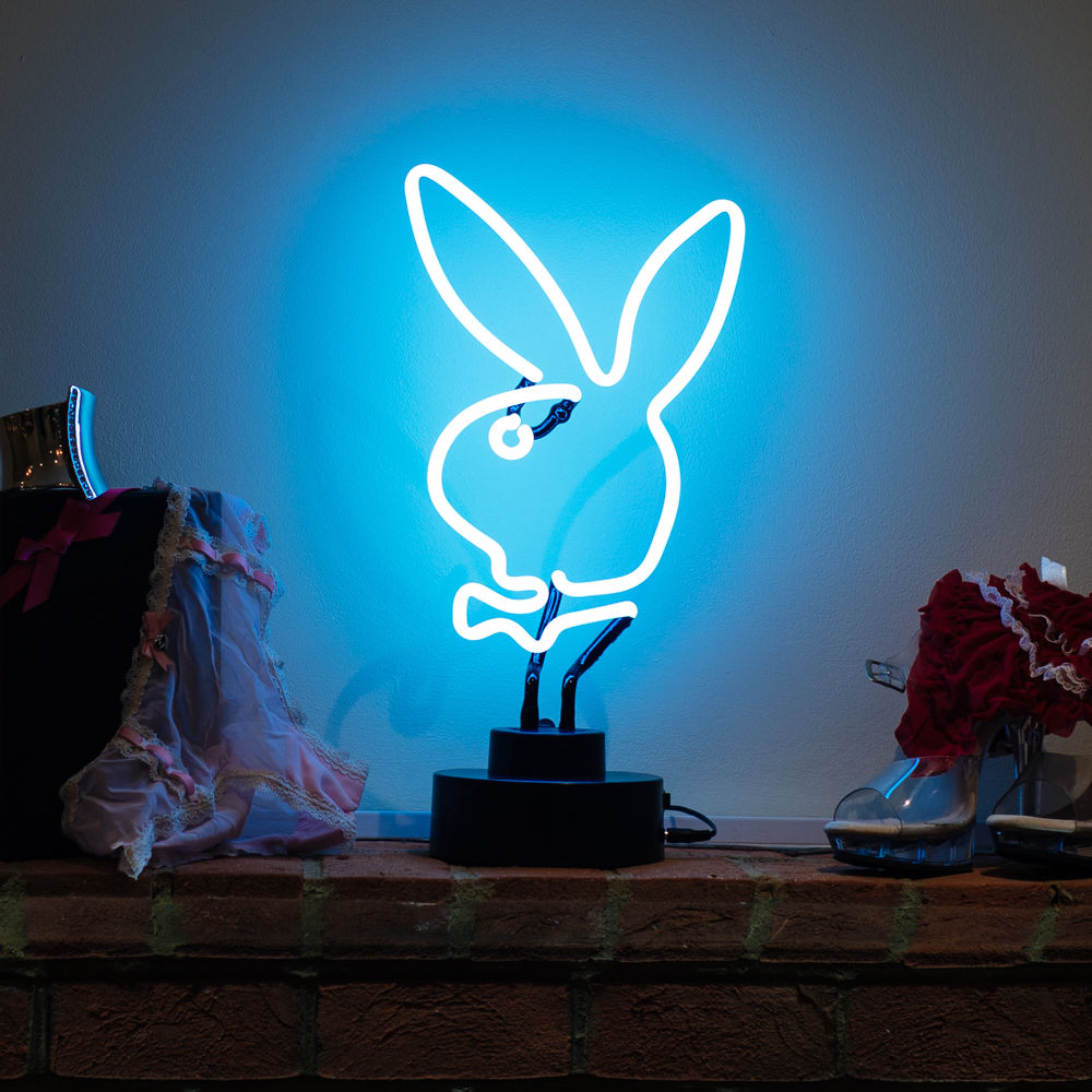 Rabbit glass table neon sign glowing red, sitting on a desk with a laptop and plants in a room, featuring visible glass tubes and wiring.