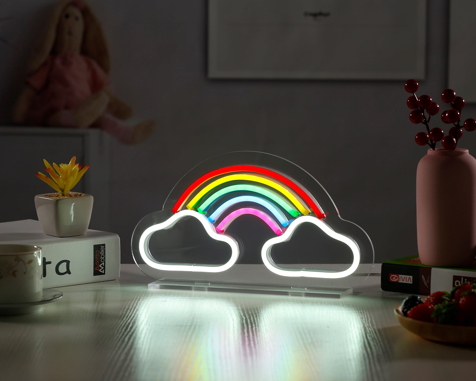 A vibrant 'Rainbow' desk neon sign with colorful glowing arches on a dark acrylic base, sitting illuminated on a wooden table next to a small plant and books.