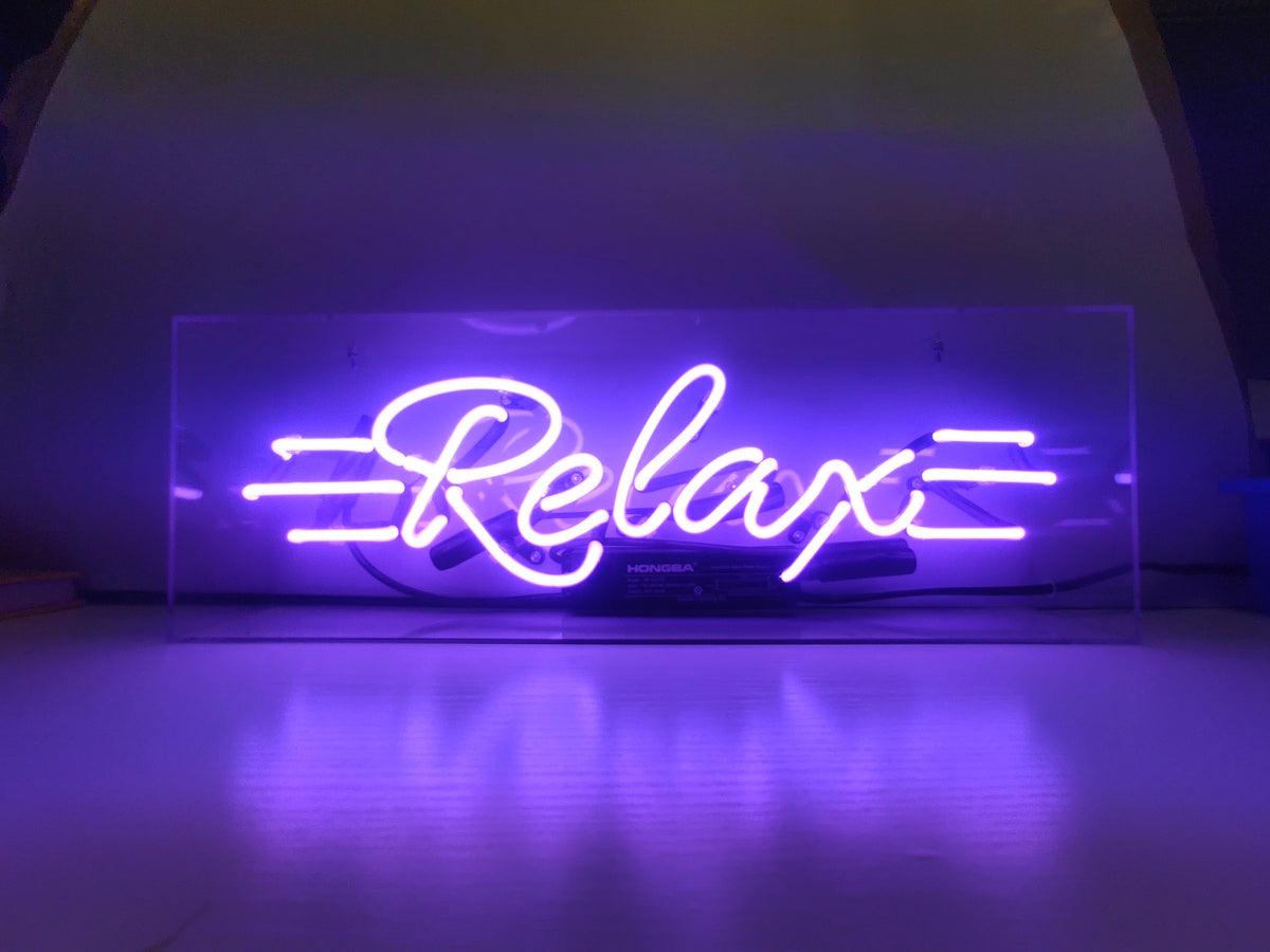 Handmade 'Relax' neon sign glowing pink inside a clear acrylic box, placed on a surface showing power details