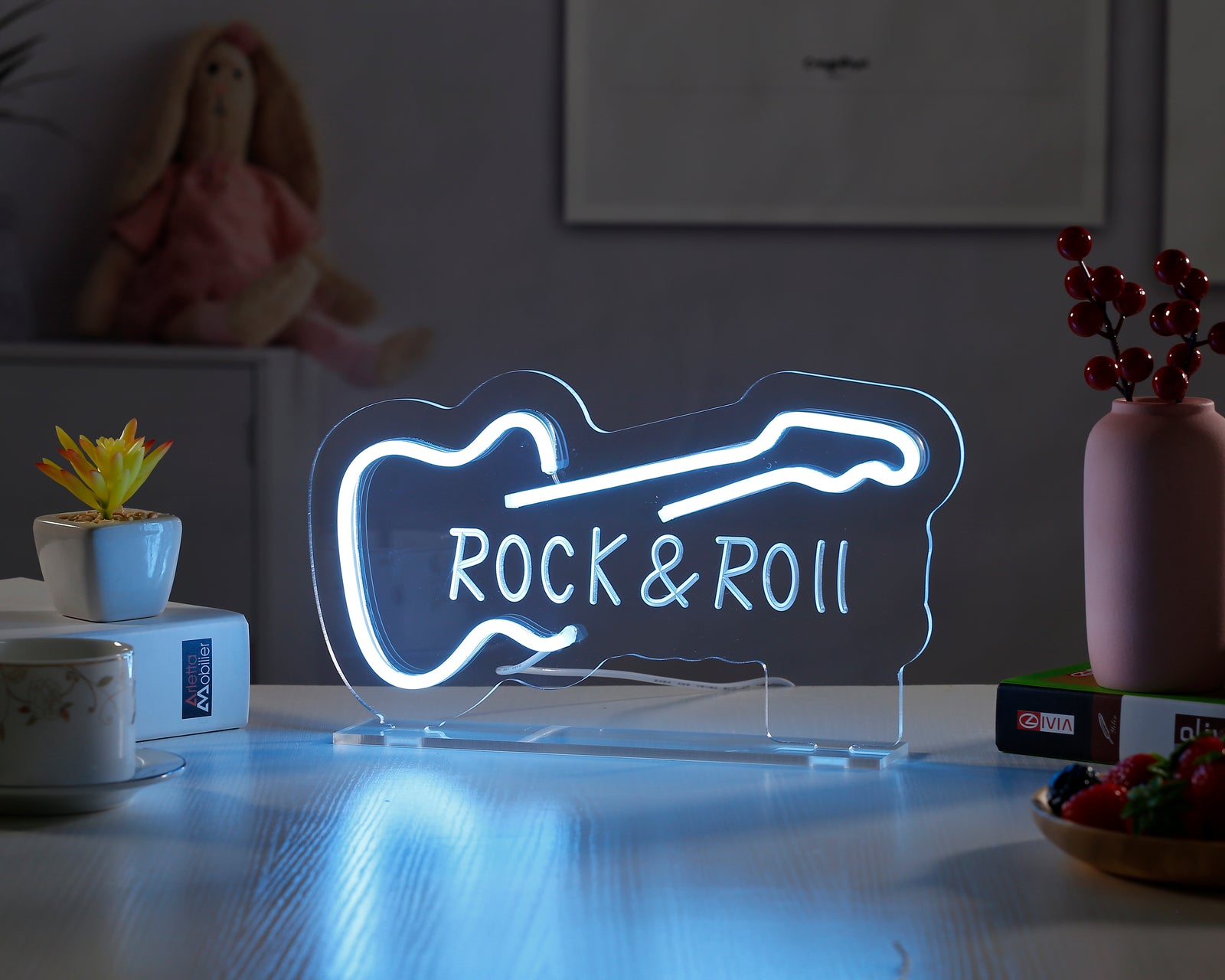 A vibrant blue and red "Rock Roll" neon desk sign displaying the rock hand gesture and lightning bolt, glowing on a shelf next to books and a plush figure.