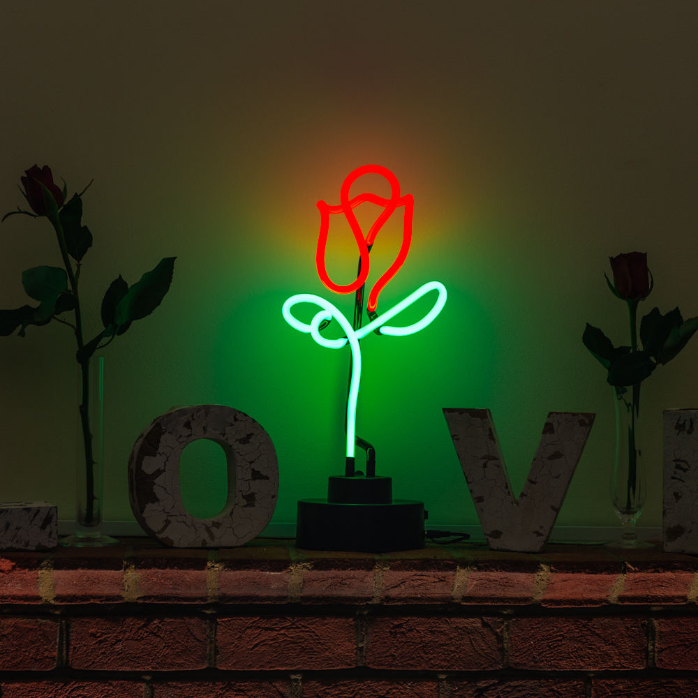 A lit neon sign depicting a red rose with a green stem and leaf, sitting on a dark wooden surface in front of blurred decorative letters and a rustic brick wall.