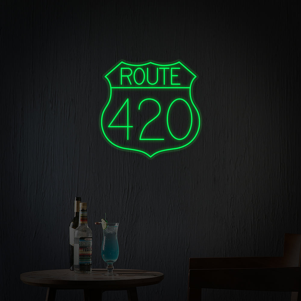 A vibrant 'Route 420 US Highway' neon sign, styled after a classic U.S. route shield in green and yellow, glows brightly against a dark, moody background featuring indistinct elements of a retro bar or hangout spot. The neon light casts a warm sheen on the wall behind it.