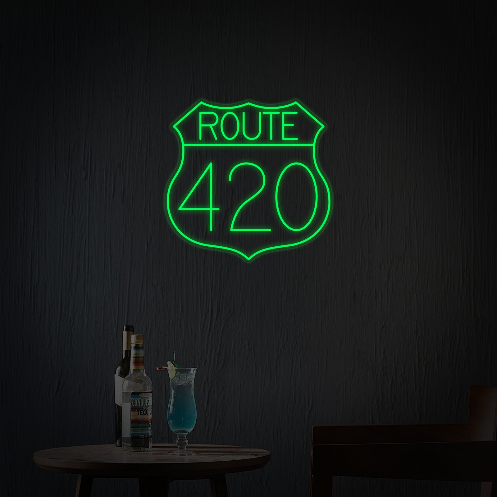 A vibrant 'Route 420 US Highway' neon sign, styled after a classic U.S. route shield in green and yellow, glows brightly against a dark, moody background featuring indistinct elements of a retro bar or hangout spot. The neon light casts a warm sheen on the wall behind it.