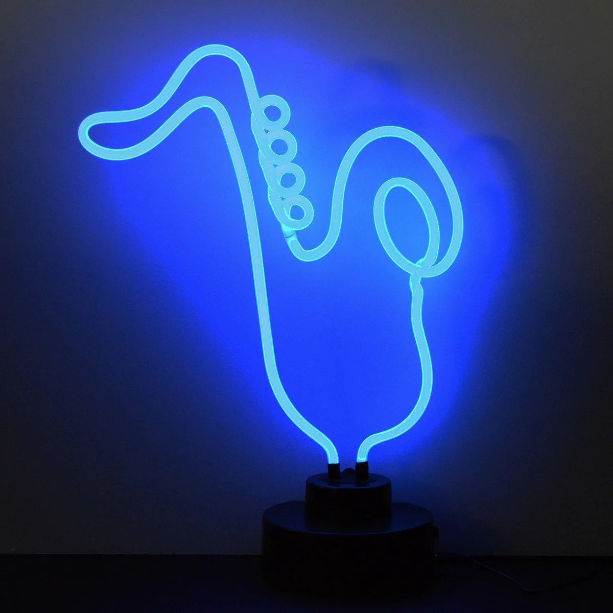 A glowing yellow neon sign shaped like a saxophone, mounted on a sleek black rectangular base on a wooden desk, against a plain beige wall.