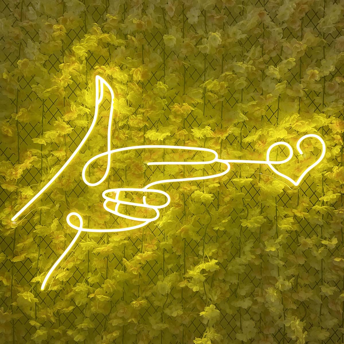 Detailed view of the 'Shoot Love Heart' neon sign illuminating against a vibrant yellow flower wall, highlighting fabrication details.
