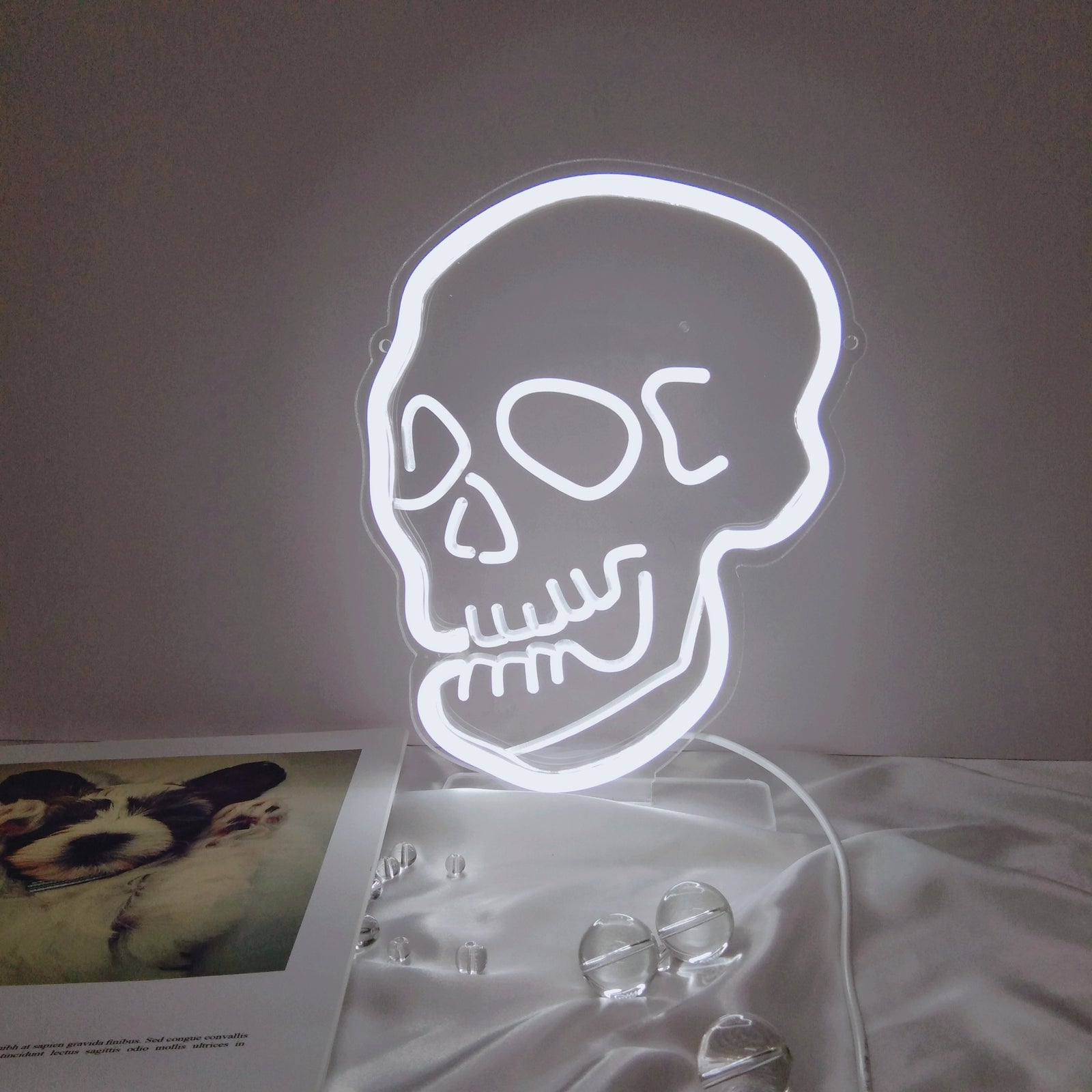 A physical, illuminated white LED neon skull sign with a clear acrylic backing, sitting on a table with a satin fabric and small beads.