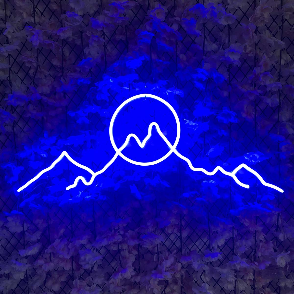 The 'Sun Mountains' neon sign glowing brightly on a wall covered in lush green artificial foliage and colorful orange and yellow flowers.