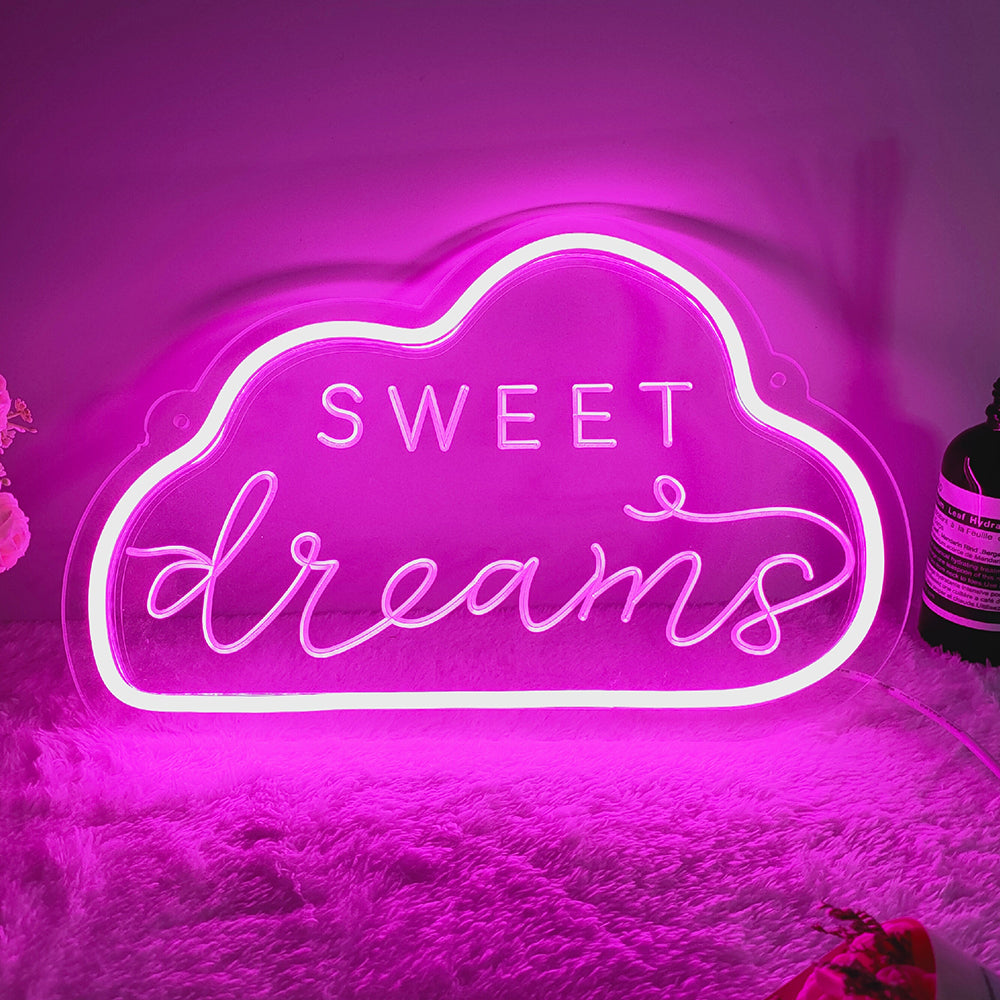 A glowing blue 'Sweet Dreams' mini neon sign mounted above a fluffy white bedspread and pillows with some floral decor nearby.