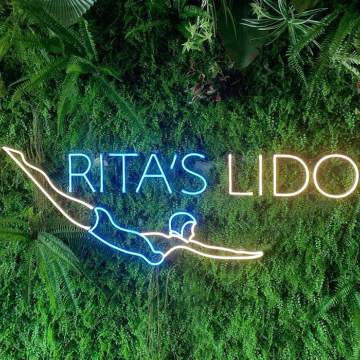 A glowing 'Swimmer Swimming Diving Club Lido' neon sign featuring a dynamic blue silhouette of a person diving, mounted on a wall covered with green artificial foliage.
