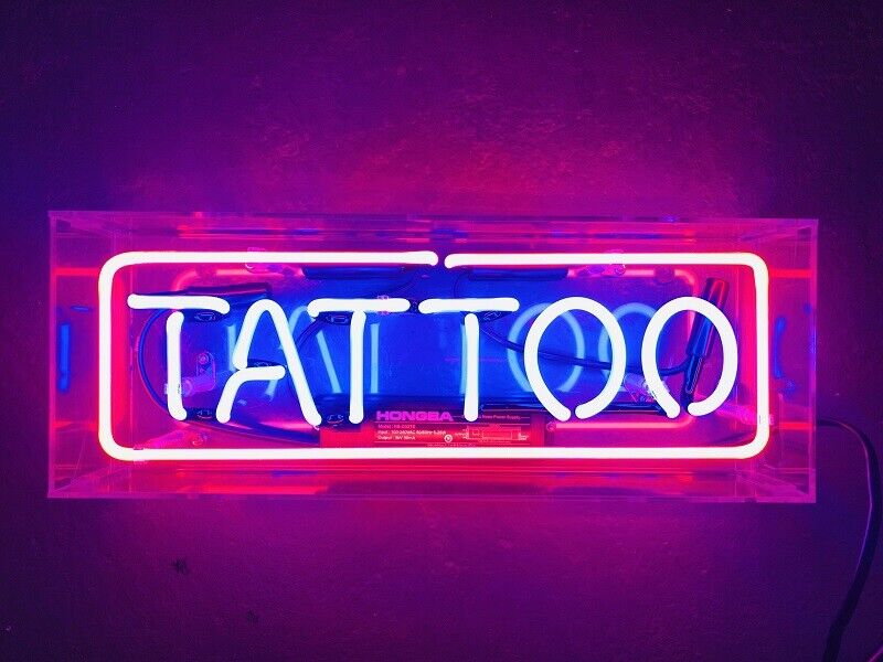 A glowing red and blue neon sign spelling "Tattoo" housed in a clear acrylic box, shown in a lit indoor setting.