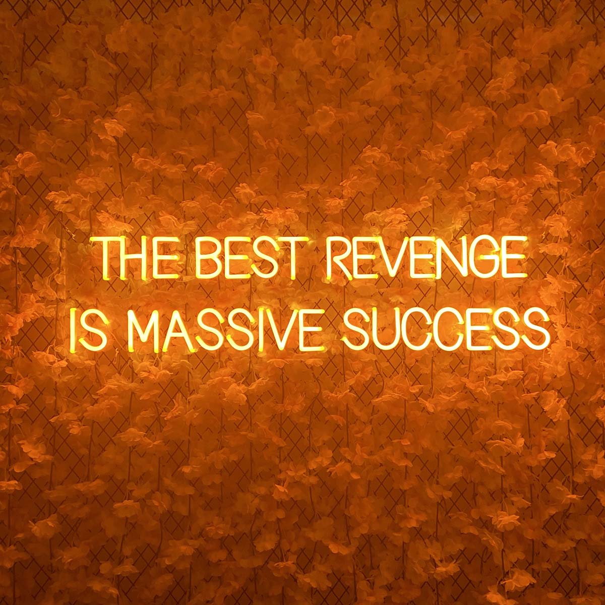 Glowing neon sign featuring the quote 'The Best Revenge is Massive Success' mounted on a wall of white flowers.
