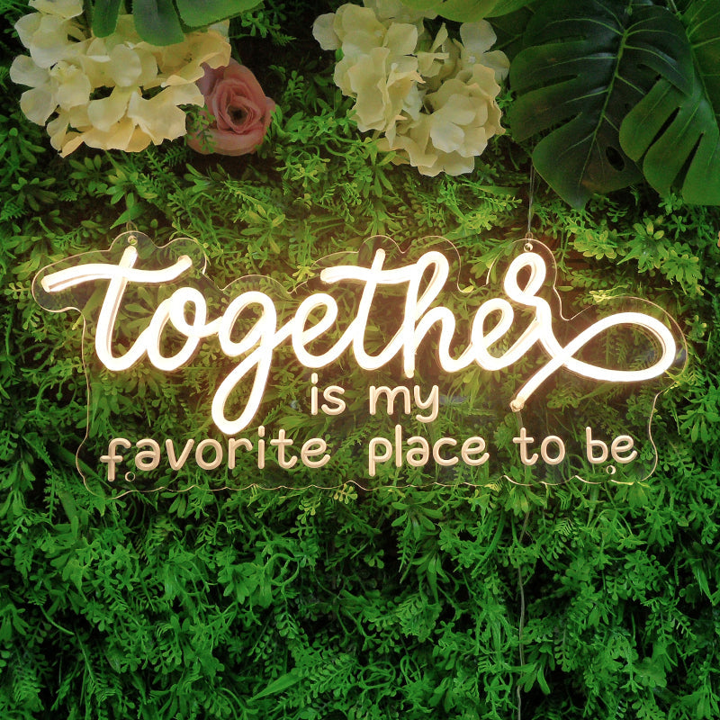 White neon sign featuring the handwritten quote 'Together Is My Favorite Place To Be' glowing brightly against a lush green wall.