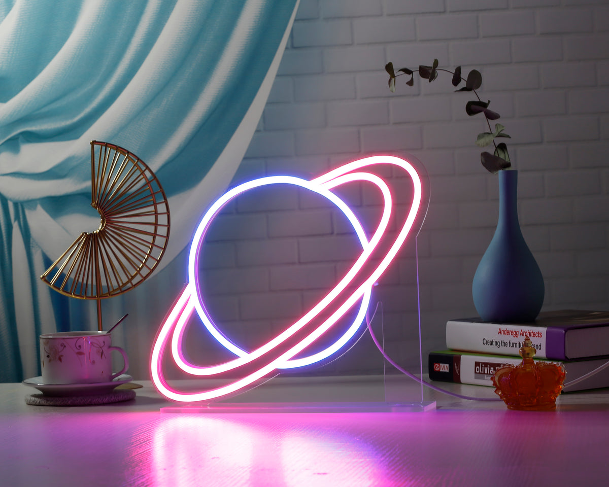 Illuminated 'Universe' desk neon sign on a dark desk next to a monitor and keyboard, featuring a green and pink alien spaceship design.