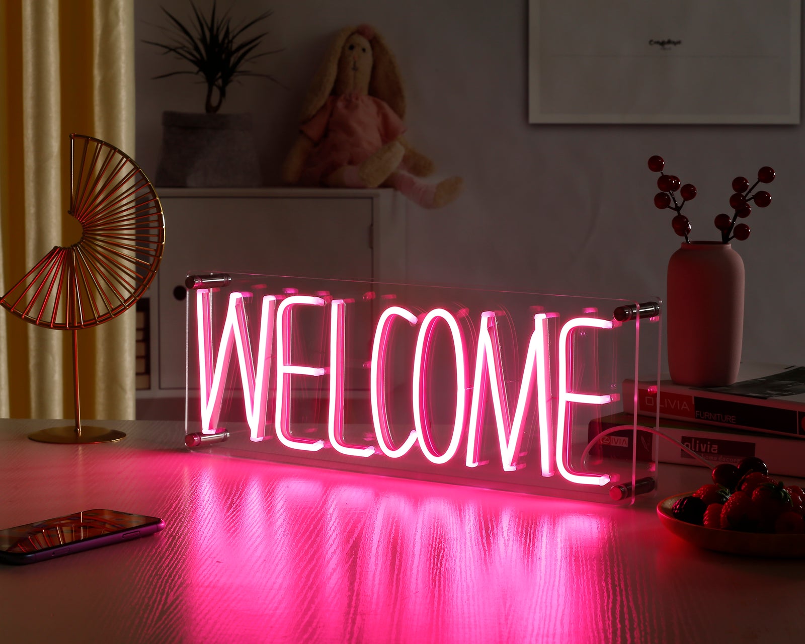 A illuminated 'Welcome' neon sign glowing with a warm pink light on a dark surface, showing the detailed letterforms and a power cable.