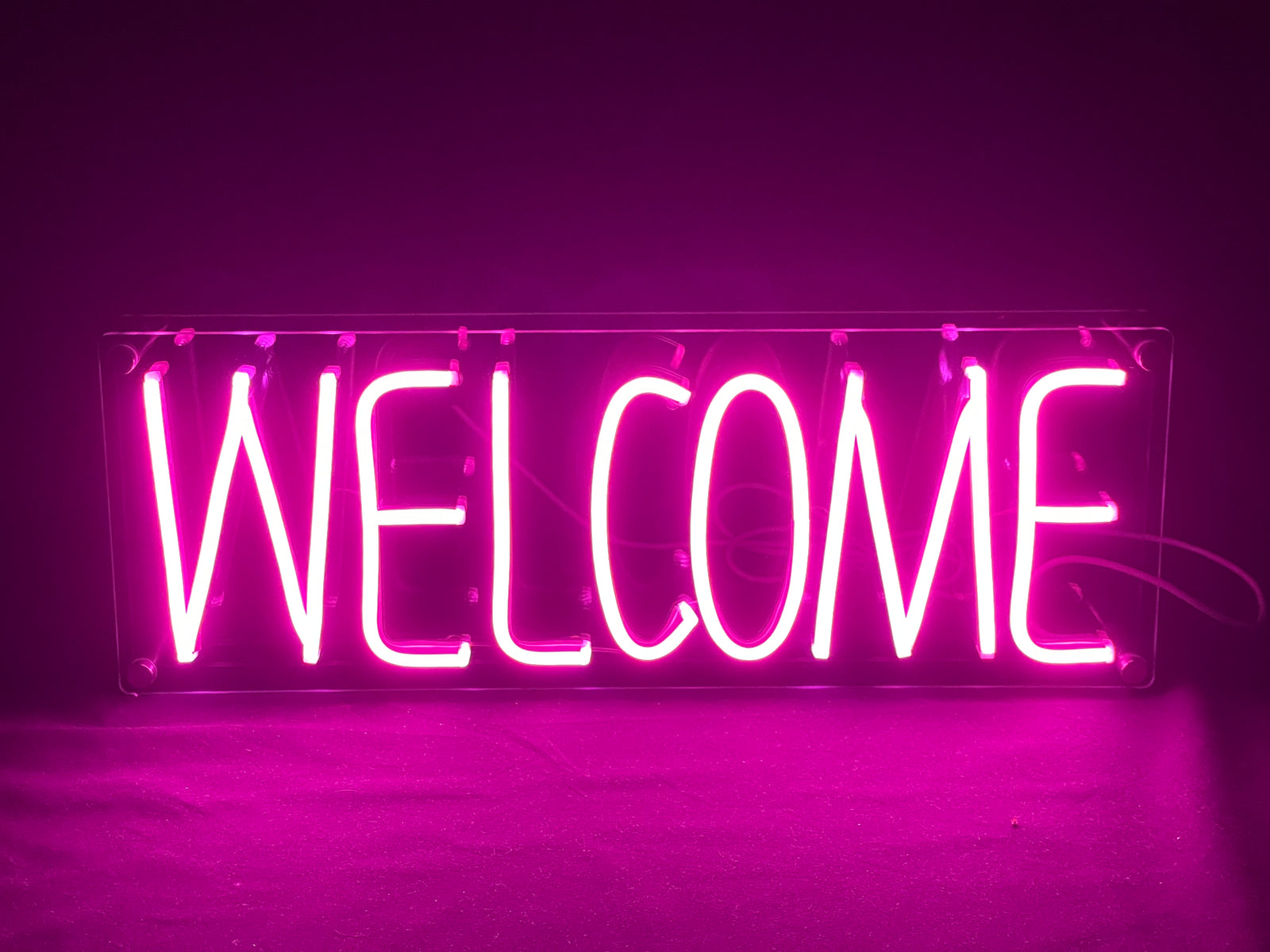 The glowing pink 'Welcome' neon sign viewed straight-on, highlighting the clear script and a slight reflected light beneath the sign.